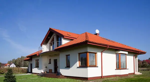 Biały dom z czerwonym dachem i balkonem na tle błękitnego nieba.